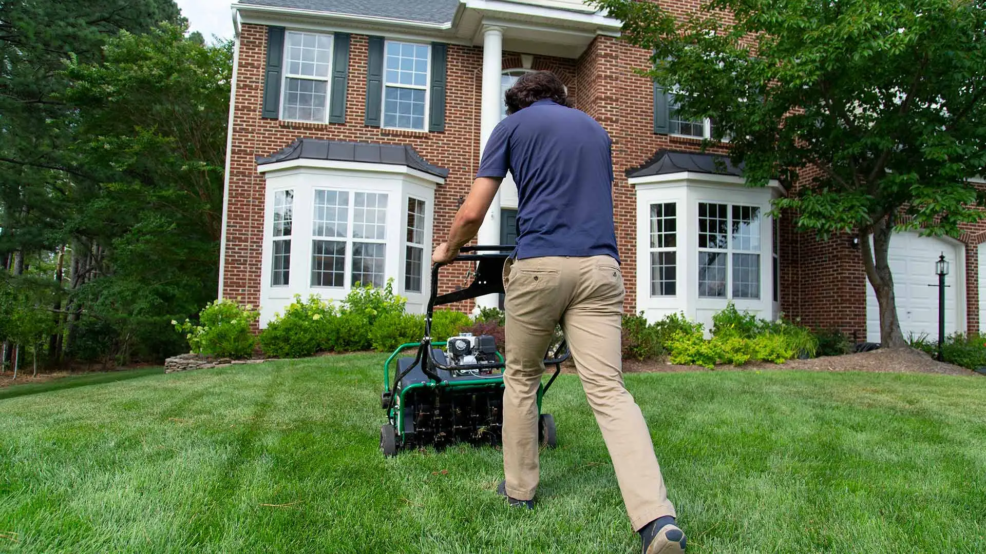 Man using an aeration machine to aerate a lawn with a home in the background.